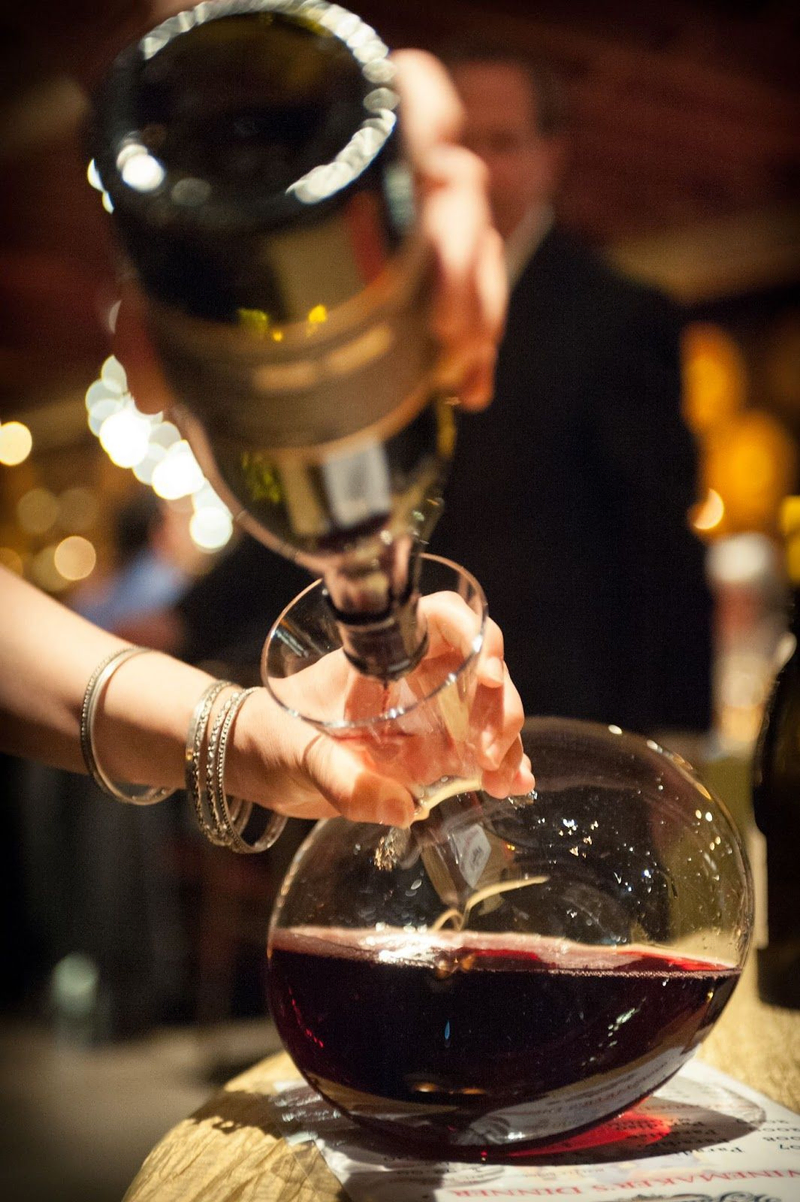 Hand pouring red wine from a bottle into a glass decanter.