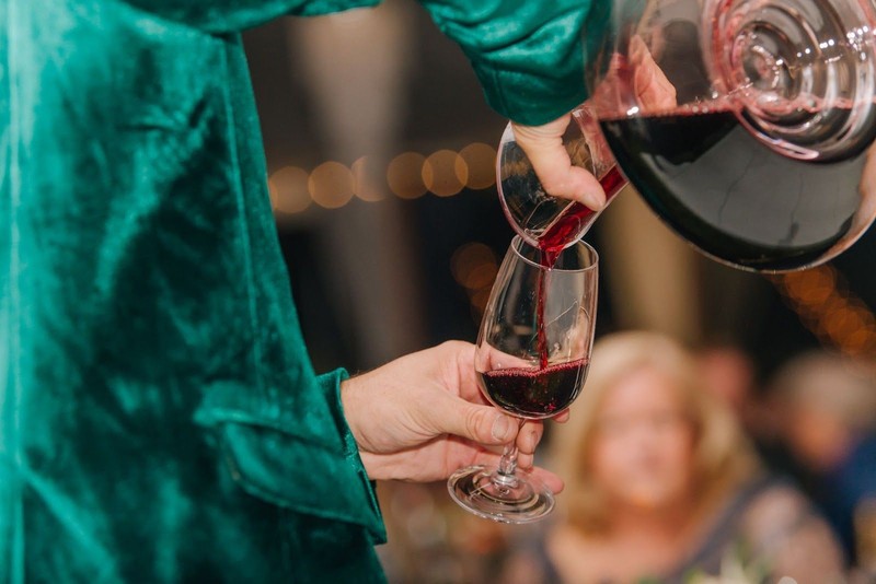 Person in green velvet pouring red wine from a decanter into a glass.