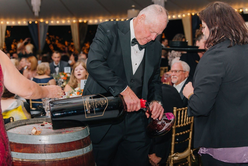 Man in a tuxedo pouring red wine from a large bottle at an event.